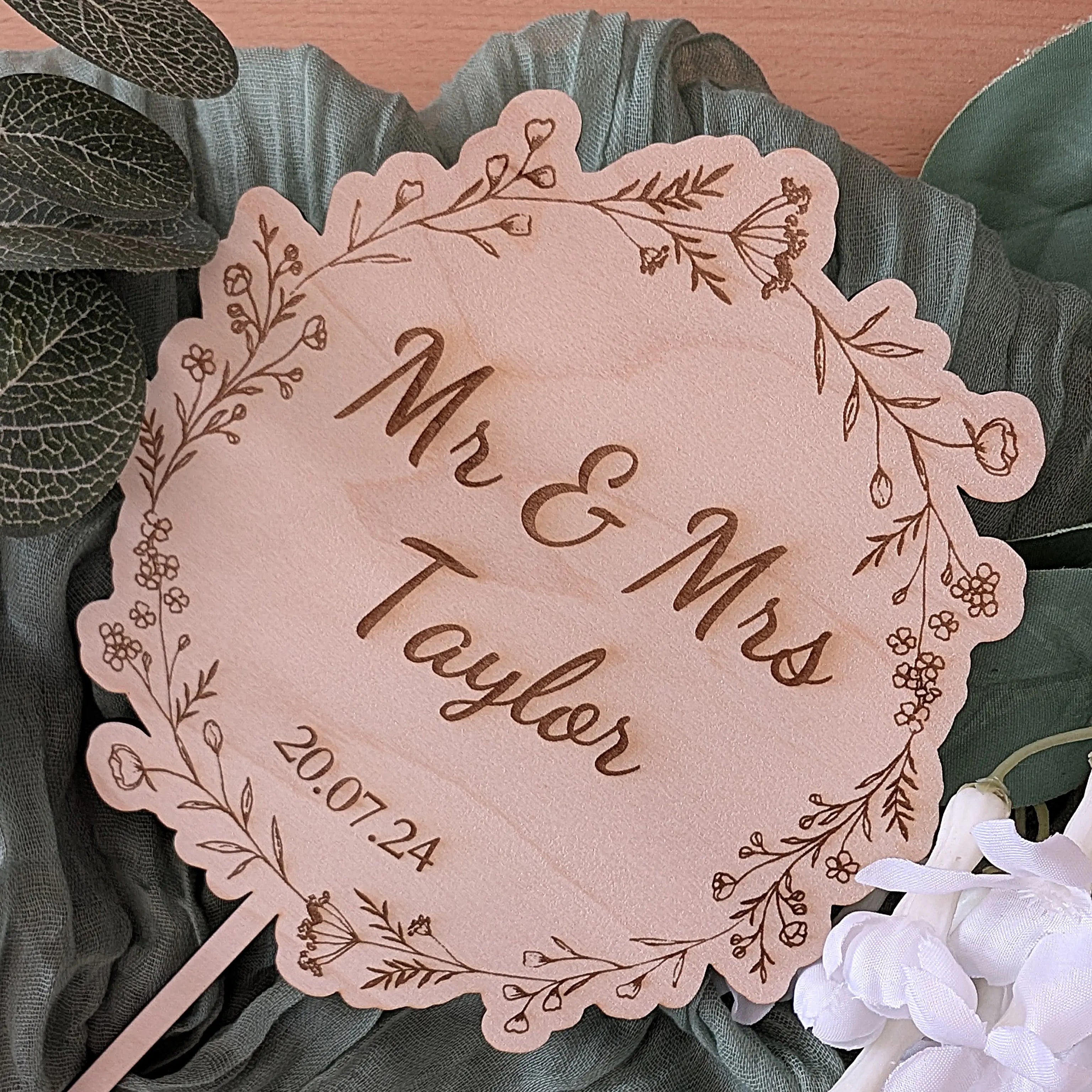 Personalized wooden plaque with 'Mr & Mrs Taylor' and date, surrounded by greenery and flowers.