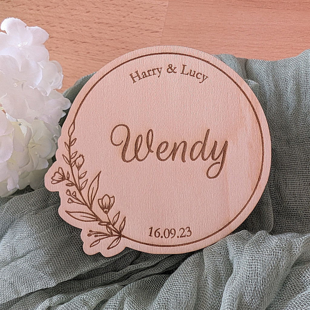 Round wooden coaster with engraved name 'Wendy' and date, placed on a fabric surface with flowers.