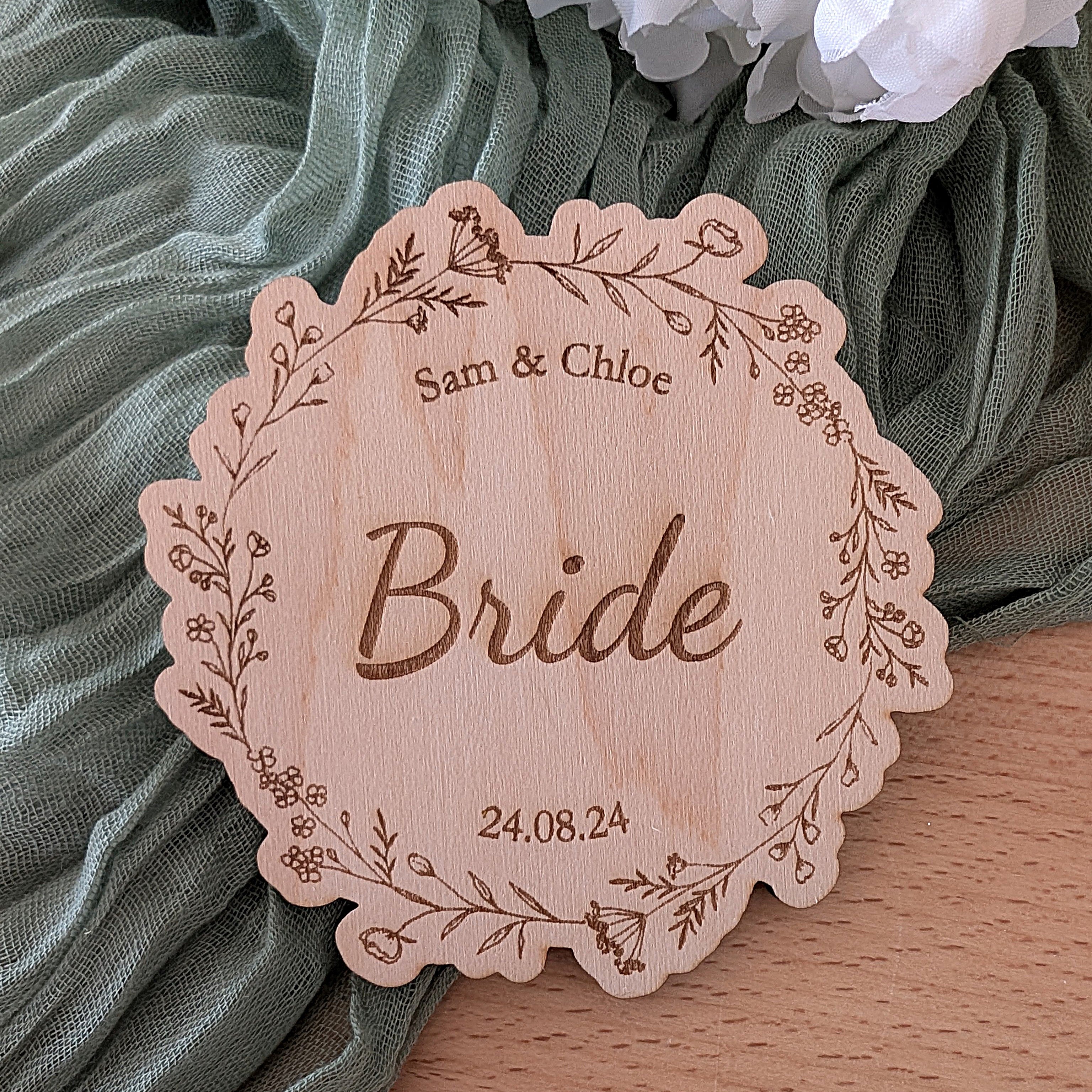 Wooden tag with 'Bride' engraved on it, surrounded by green fabric and flowers.