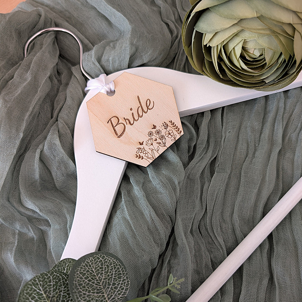 White wedding hanger with 'Bride' tag on a green fabric background along with a green rose

