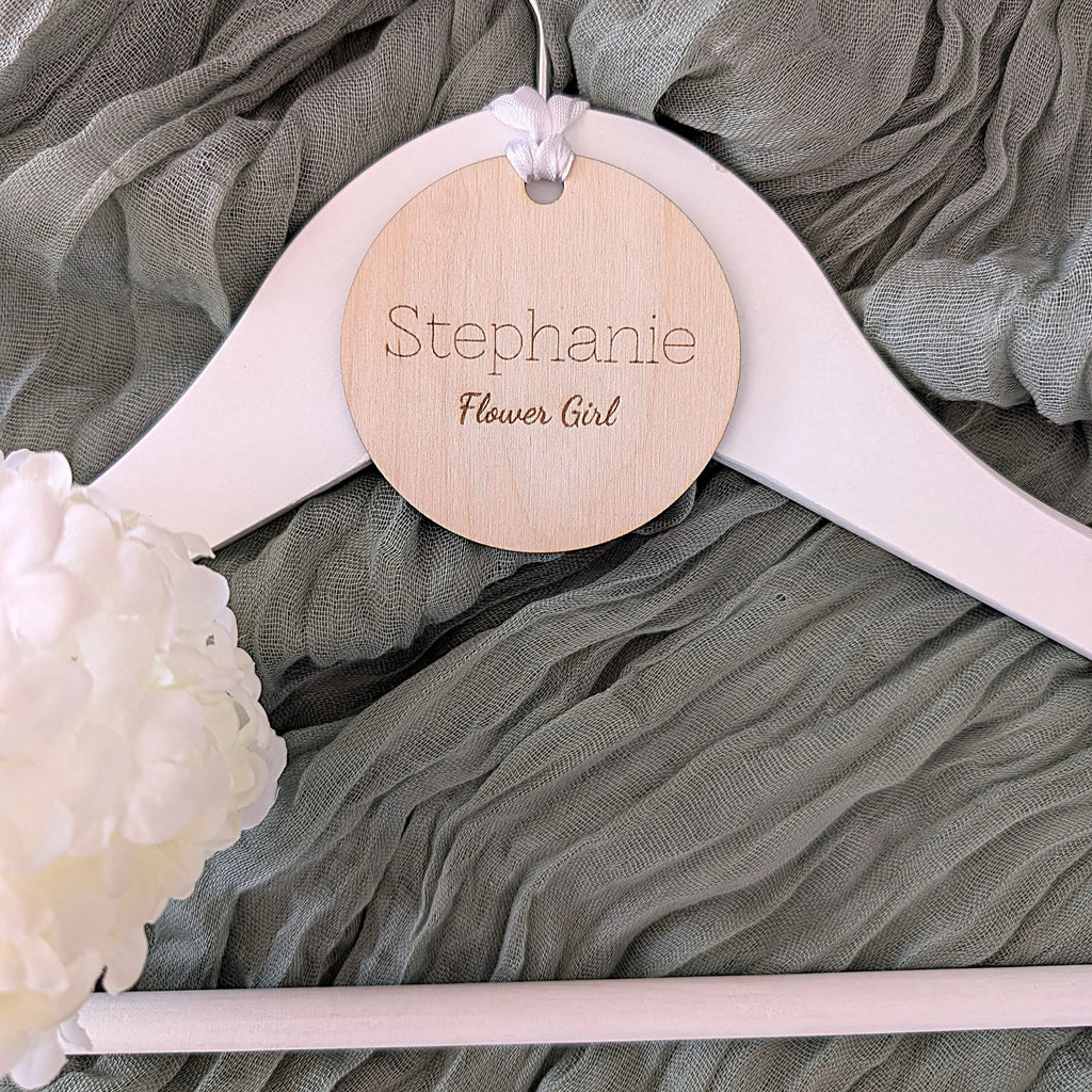 White hanger with a wooden tag labeled 'Stephanie Flower Girl' on a gray fabric background.