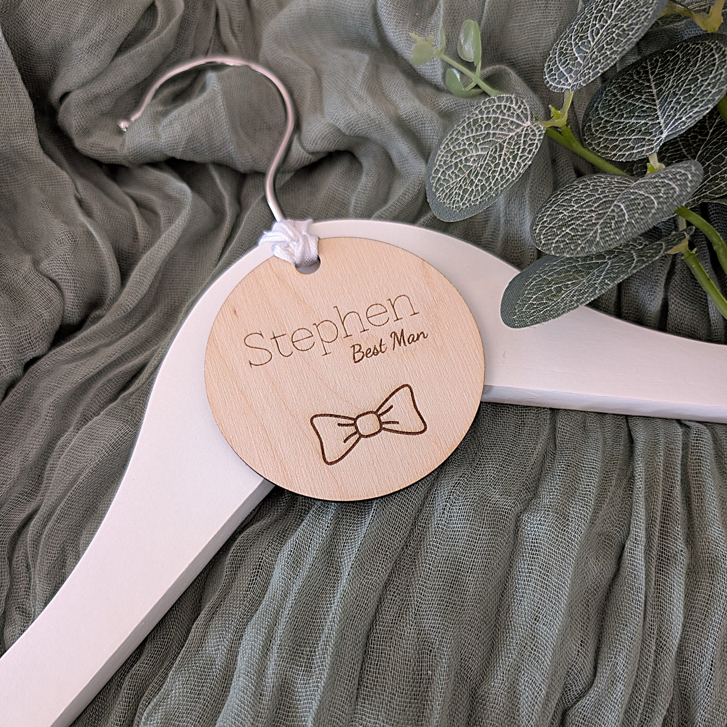 Personalized wooden tag on a hanger with 'Stephen Best Man' text and bow tie icon, against a green fabric background.