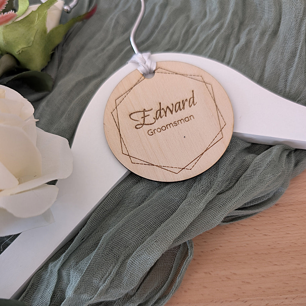 Wooden tag on a hanger with 'Edward Groomsman' text, surrounded by flowers and fabric.