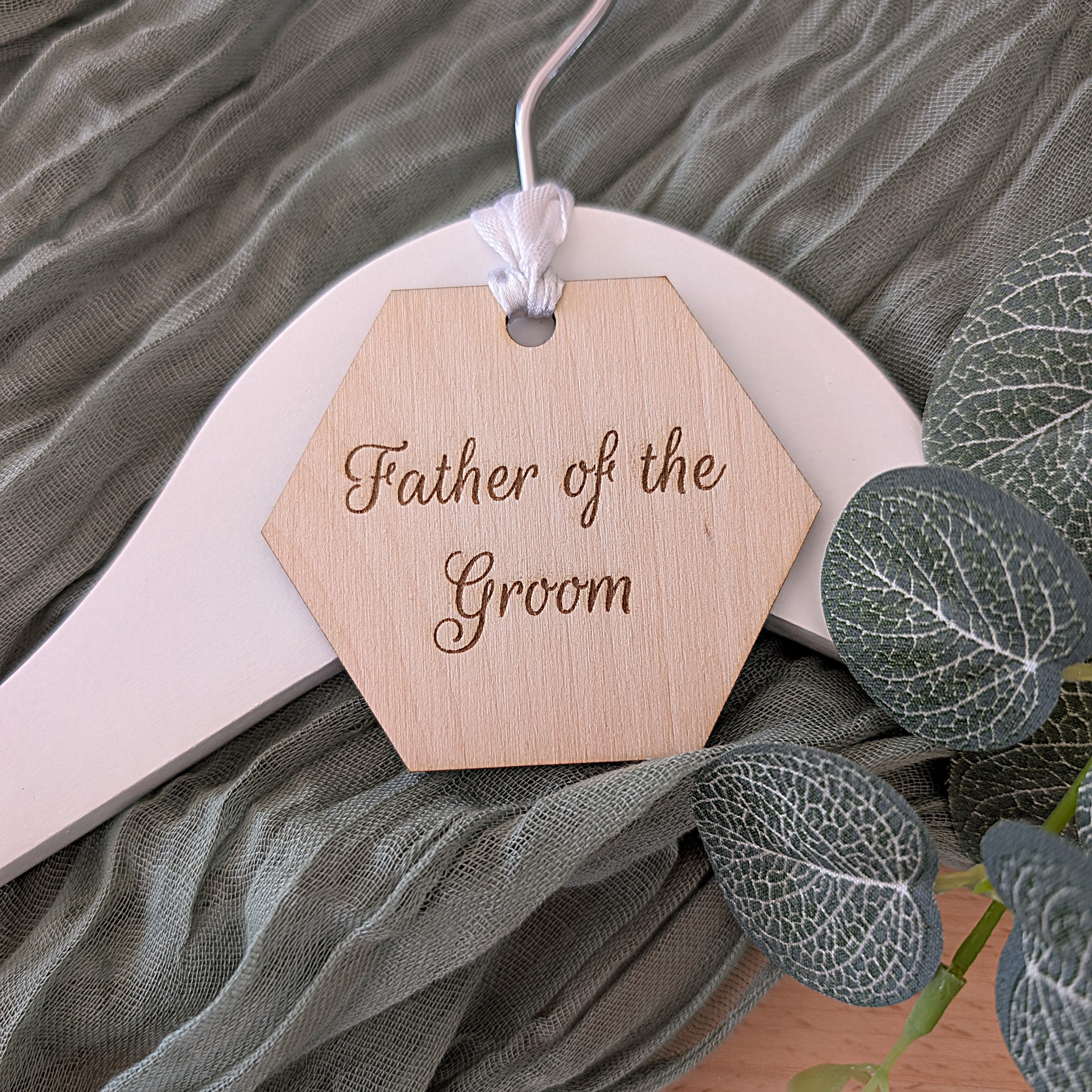 Wooden tag with 'Father of the Groom' text on a hanger with green fabric and leaves in the background