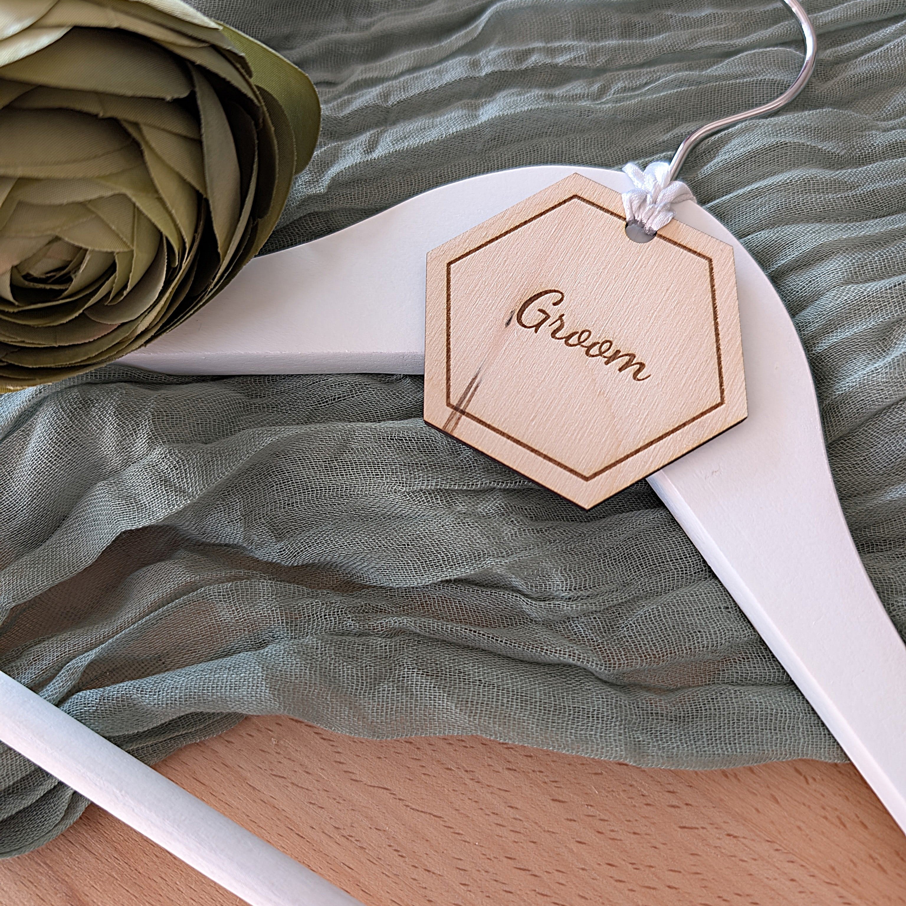 Hexagonal wooden tag labeled 'Groom' on a white hanger with green fabric and flower in the background.