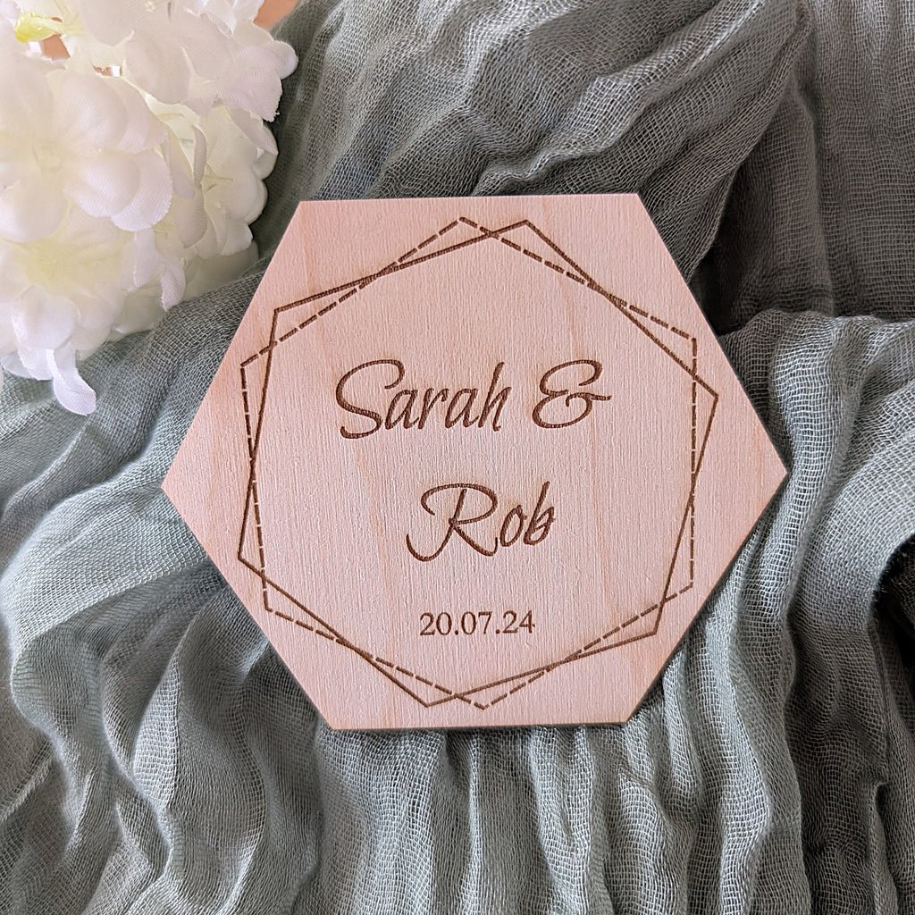 Hexagonal wooden coaster with engraved names 'Sarah & Rob' and date '20.07.24' on a soft fabric background.